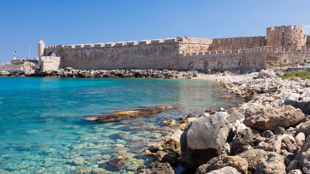 Μεσαιωνικά τείχη στη Ρόδο δίπλα σε καταγάλανα νερά, συνδυάζοντας ιστορία και φυσική ομορφιά.