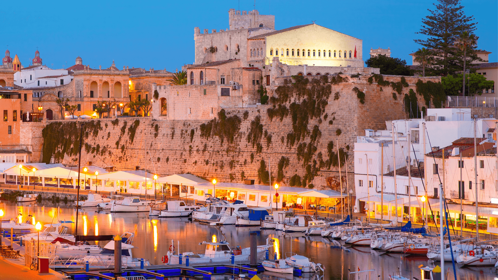 La ville de Ciutadella de Menorca, en soirée, avec les lumières éclairant l'eau transparente du petit port.