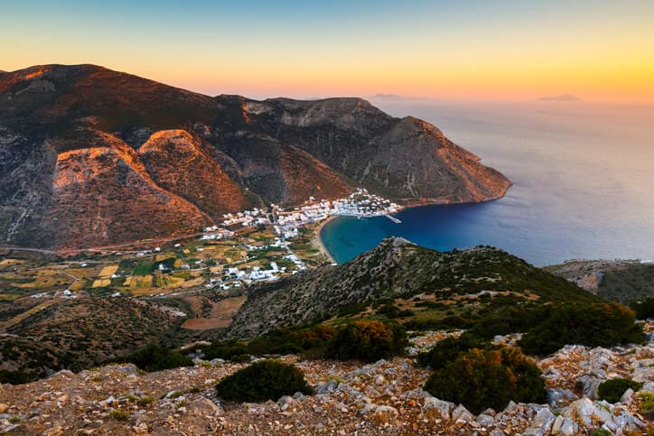 Vista panoramica di Folegandros al tramonto con il villaggio costiero, la baia e le montagne rocciose affacciate sul Mar Egeo.