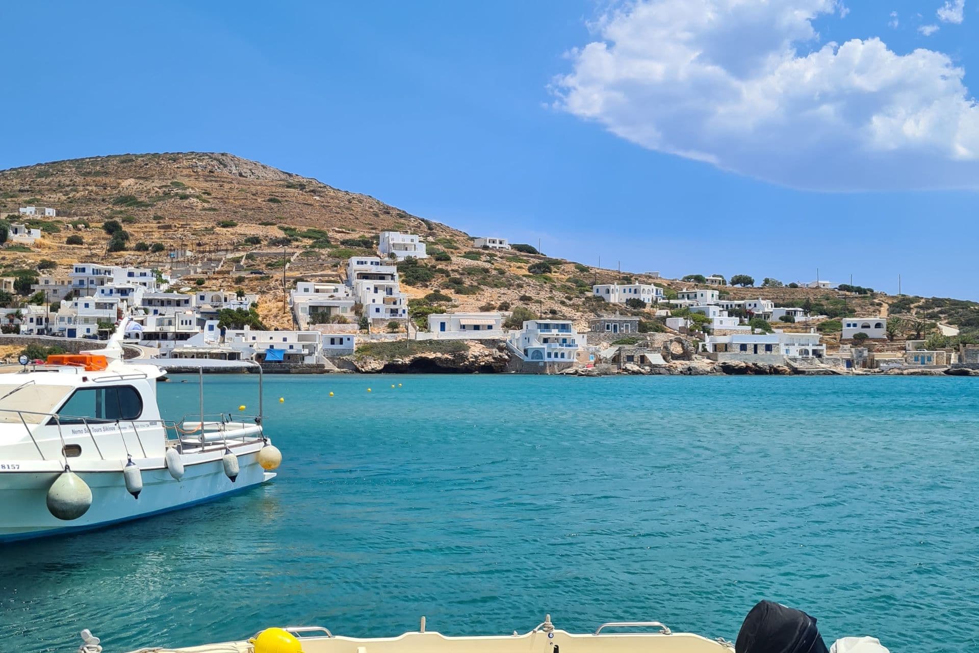 Petit port avec un bateau amarré sur Sikinos, avec en toile de fond une colline et des petites maisons blanches.