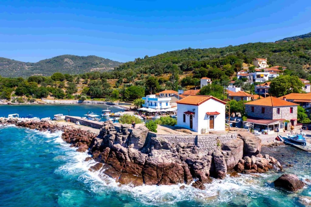 Ένα γραφικό παραθαλάσσιο σκηνικό στη Λέσβο, όπου τα πέτρινα σπίτια με τα κεραμίδια αγκαλιάζουν το λιμάνι και τα καταγάλανα νερά του Αιγαίου, αποτυπώνοντας τη γαλήνια ομορφιά του νησιού.