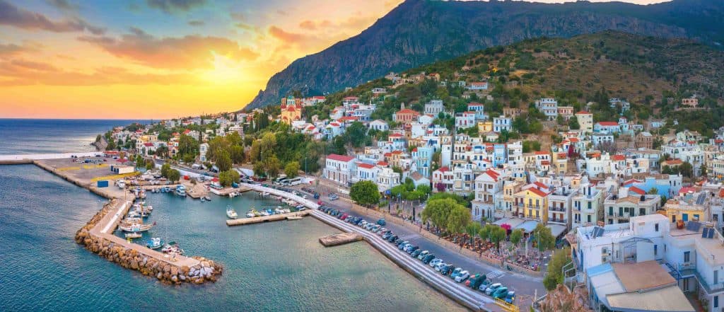 Πανοραμική θέα σε παραθαλάσσιο οικισμό της Ικαρίας στο ηλιοβασίλεμα, με το λιμάνι και τα πολύχρωμα σπίτια να αγκαλιάζουν το Αιγαίο.