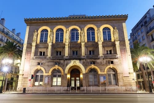 Classical building with arched doors and sculpted figures illuminated at twilight on an empty city street with palm trees.
