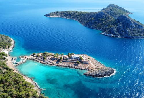 Aerial view of a serene Agistri island, showcasing clear turquoise waters, a rugged coastline, and lush greenery under a bright blue sky.