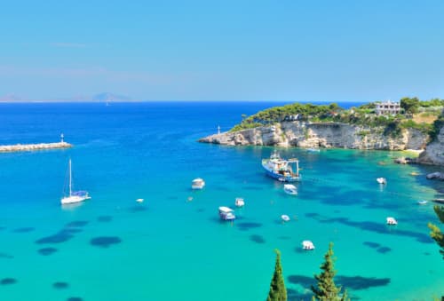 Crystal-clear waters and moored boats off the rocky coast of Alonissos, with lush greenery and a clear blue sky enhancing the tranquil scene.