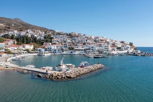 Calm waters of a small marina in Andros, Greece, with white-washed houses climbing the hillside, fishing boats docked, and a clear sky overhead.