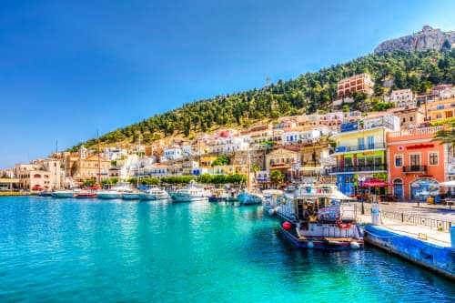 Yachts docked in a vibrant waterfront with multicolored buildings ascending a hill, beneath a clear blue sky in Kalymnos, Greece.