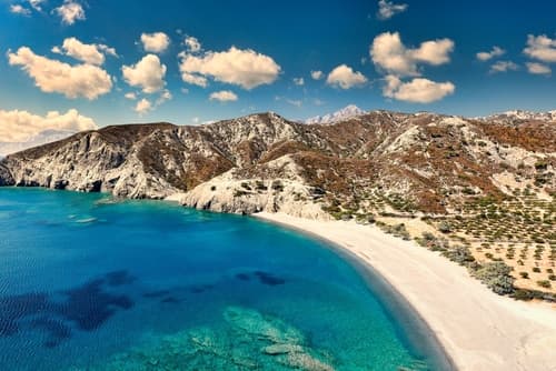 Vue aérienne d'une plage de sable blanc incurvée aux eaux claires et turquoises, bordée de montagnes escarpées sous un ciel parsemé de nuages sur une île grecque.