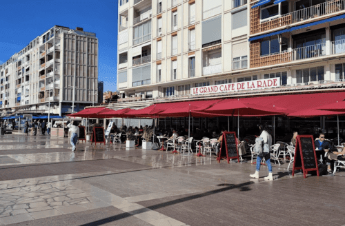 Spacious outdoor seating at "LE GRAND CAFÉ DE LA RADE" with red awnings, on a bustling promenade under a clear blue sky.