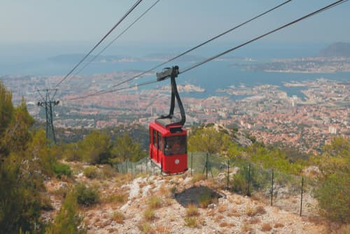 Red cable car ascending over a hilly landscape with a panoramic view of a coastal city and blue sea in the background.