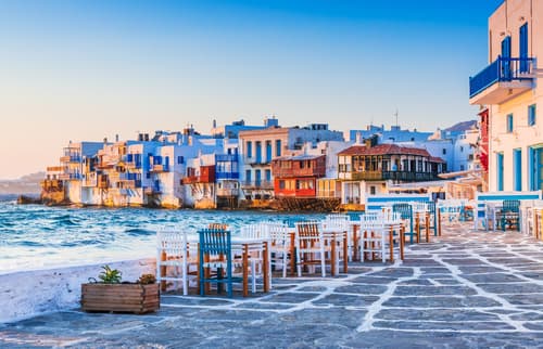 Seaside dining area in Mykonos, Greece, with empty tables and chairs facing the colourful houses on the waterfront bathed in the warm glow of sunset.