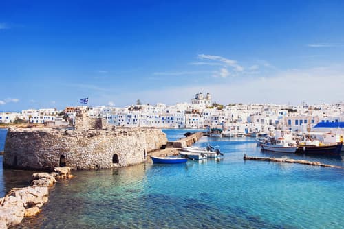 Harbour view in Paros, Greece, featuring traditional Cycladic architecture, boats moored in clear blue water, and remnants of an old fort under a bright blue sky.
