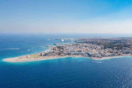Aerial view of a coastal town with a curving beachfront, clear blue waters, and dense urban development on a sunny day, in Rhodes, Greece.