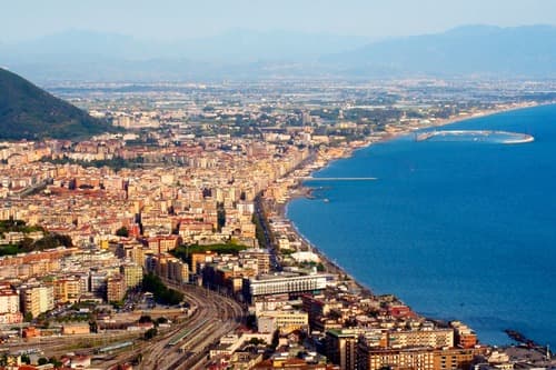 Aerial view of Salerno’s coastal cityscape, with dense buildings stretching along the curving shoreline of the Tyrrhenian Sea, backed by rolling hills.