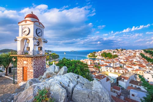 Iconic clock tower overlooking the terracotta rooftops of Skiathos town, with the azure sea in the background under a bright, cloud-streaked sky.