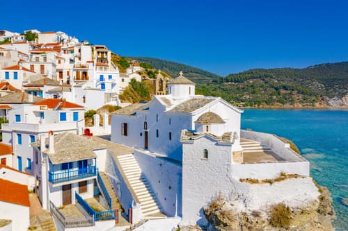 Whitewashed buildings with blue accents cascade down the hillside to the sea in Skopelos, showcasing traditional Greek island architecture under a vibrant sky.