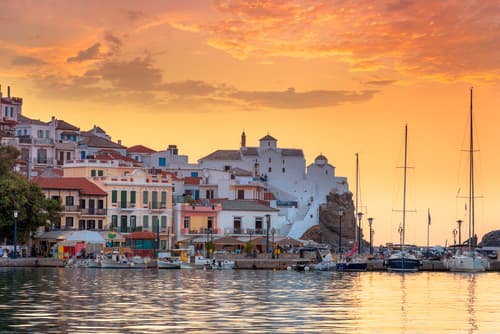 A tranquil harbor with moored yachts against a backdrop of Skopelos town's terraced buildings, under a vibrant sunset sky.