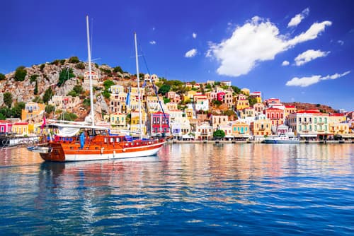 Boats moored in a harbor with colorful buildings on the hillside under a blue sky with fluffy clouds, in Symi Island, Greece.