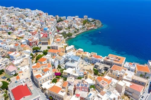 Aerial view of Syros island, Greece, with densely packed traditional white houses, a prominent church dome, and azure waters along the coastline.