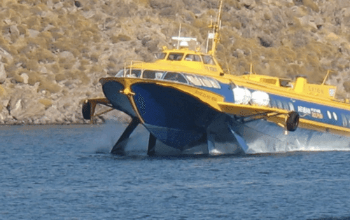 Yellow and blue hydrofoil vessel of Aegean Flying Dolphins approaches a rocky coast, its bow raised above water, ready to dock.