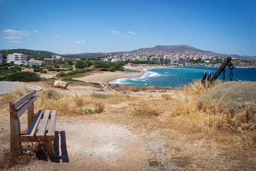 Coastal landscape of Rafina, Greece, with a lone bench overlooking a dry grassy field, sandy beach, and a view of the town against a mountain backdrop.