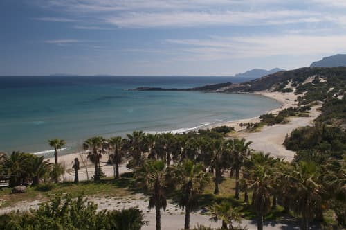 Deserted sandy beach with palm trees leading to turquoise waters, under a hazy sky, nestled between rolling hills on a peaceful Greek isle.