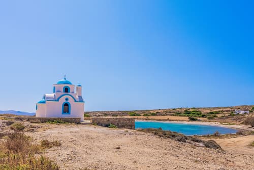 A traditional blue-domed Greek church stands alone in a dry landscape, overlooking a tranquil bay on the sun-drenched island of Lipsi.