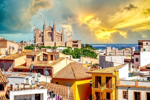 An image of a historic cathedral rising above terracotta-roofed houses against a dramatic golden sunset sky with clouds, viewed from an urban vantage point.