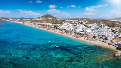 Aerial view of a coastline in Naxos, Greece, with crystal clear turquoise waters, white buildings, sandy beach, and a lush hill under a sunny sky.