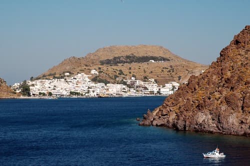 A small boat sails near the rocky coastline with a view of a hillside Greek village cascading down to the sea under a clear blue sky.