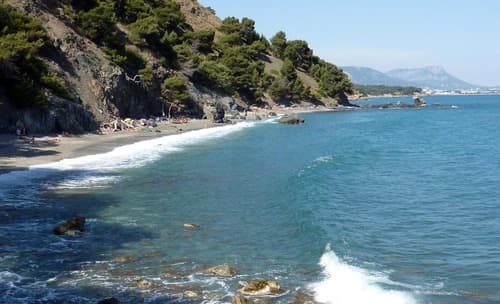 Secluded pebble beach with clear blue waters, bordered by green hills, under a clear sky with distant mountains.