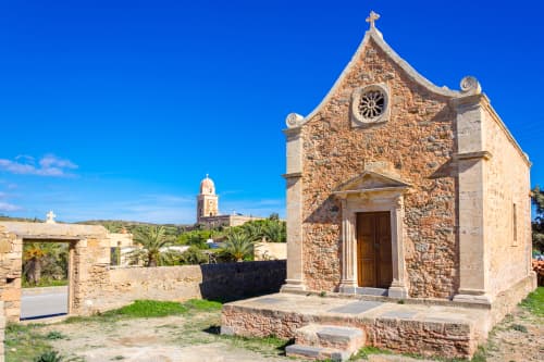 Small traditional stone church with a pointed facade under a clear blue sky, with palm trees and a bell tower in the background.