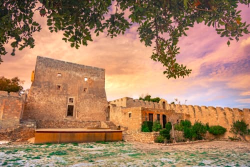 Historic stone fortress with weathered walls under a pastel sunset sky, framed by tree branches in the foreground.