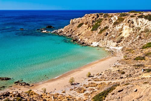 Secluded cove with turquoise waters and a sandy beach, bordered by rugged cliffs under a clear blue sky, on Crete's picturesque coast.