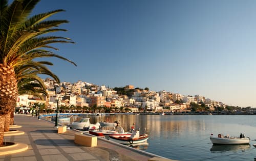 Palm-lined waterfront promenade in Sitia with moored boats, calm sea, and a backdrop of white houses against a clear sky at sunset.