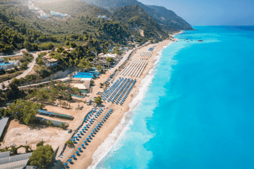 Açık mavi sulara sahip kumlu plaj, mavi renkte şezlong ve şemsiyelerin yanında yeşil ağaçlar ve bitkilerle dolu bir alan