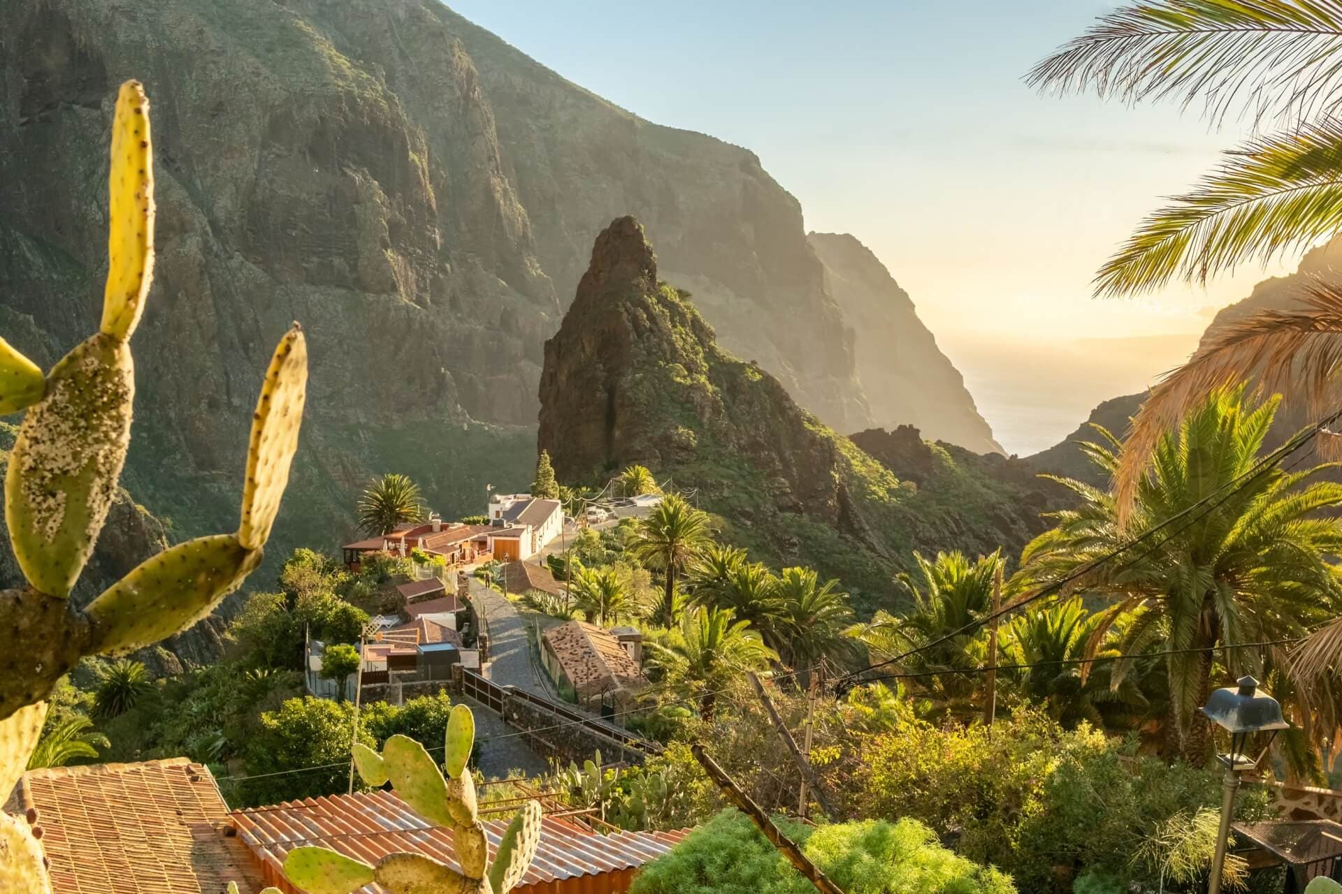 A picturesque village nestled in the lush mountains of the Canary Islands, surrounded by towering rock formations, palm trees, and a serene sunset view.