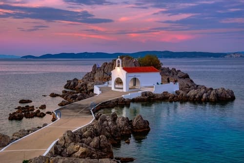 Gün batımında, sakin sularla çevrili kayalık bir çıkıntı üzerinde, kırmızı çatılı küçük beyaz bir şapel.