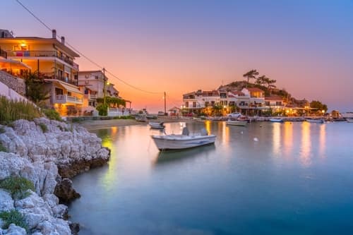 A tranquil harbour at sunset, with illuminated waterfront houses, calm waters, and a small boat reflecting the soft evening light.