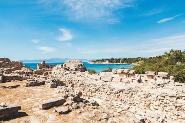 Resti archeologici di un antico sito in pietra con vista sul mare azzurro e una baia circondata da vegetazione mediterranea