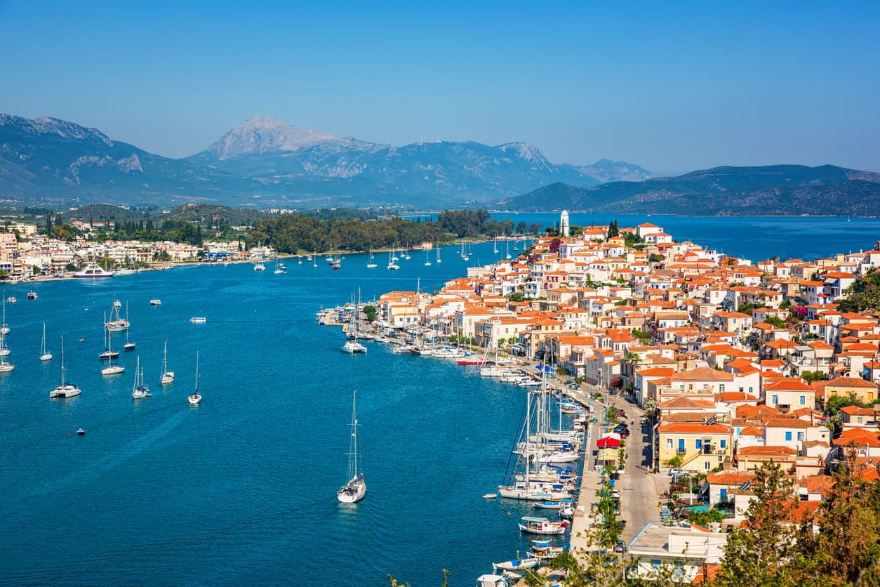 Panoramic view of Poros, with the seaside front of the town, the traditional houses and the picturesque harbor full of sailboats and boats, a breath away from the coast of Peloponnese.
