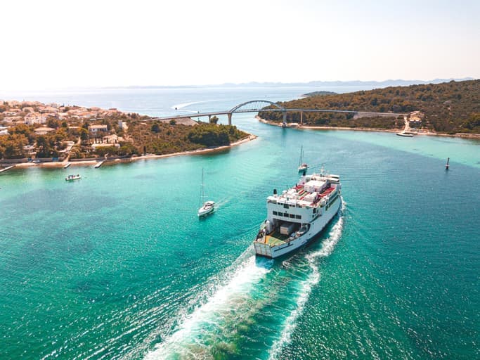 Ferry cruzando un canal turquesa rodeado de islas verdes y una calzada bajo un cielo despejado.