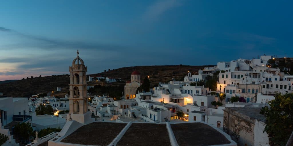 Η Τήνος λίγο πριν πέσει η νύχτα: τα κατάλευκα σπίτια φωτίζονται απαλά, το καμπαναριό στέκει αγέρωχο και ο οικισμός απλώνεται ήρεμα στην πλαγιά.