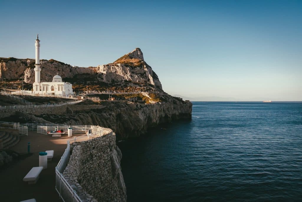 A white church on the left side sitting at the base of the rock of Gibraltar, with the sea on the right side.