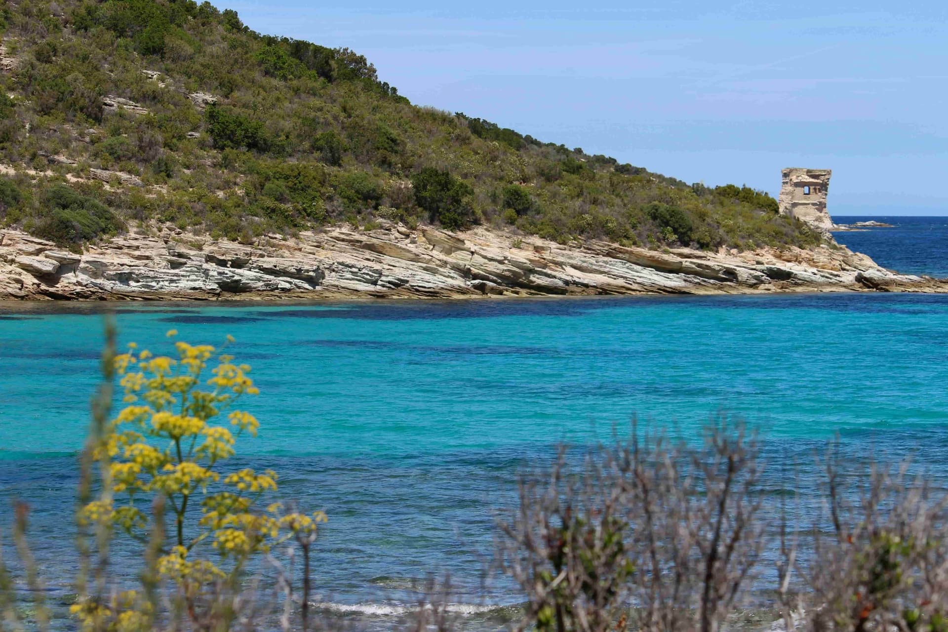 Une berge rocheuse au-dessus d'une crique azure où trône une petite tour de guet de pierre.