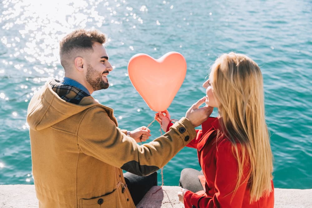 Una coppia sorridente si gode un momento romantico vicino al mare, con un palloncino a forma di cuore, simbolo perfetto di un San Valentino speciale.