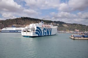 A GNV ferry entering the Port of Barcelona, with a Grimaldi Lines ferry docked in the background and cargo containers along the pier.