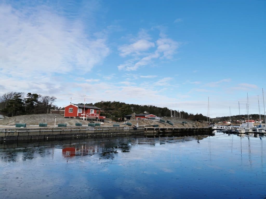 Scenic waterfront in Larvik, Norway, featuring red wooden houses, a calm harbor with moored sailboats, and a clear blue sky reflecting on the water