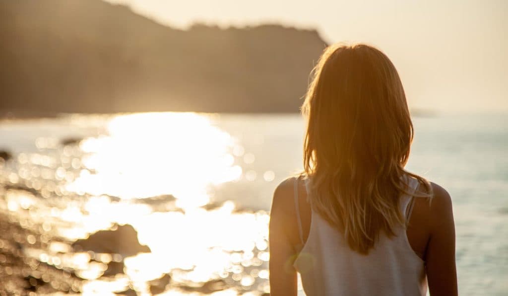 Donna di spalle in riva al mare al tramonto, mentre osserva l'acqua scintillante e la costa in lontananza.
