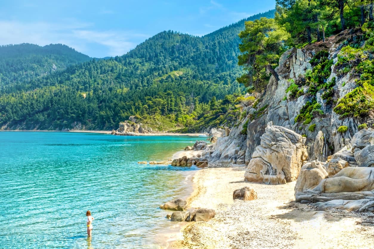 Fava Beach in Griechenland mit Felsen, Sandstrand und klarem, türkisfarbenem Wasser vor grüner Bergkulisse.